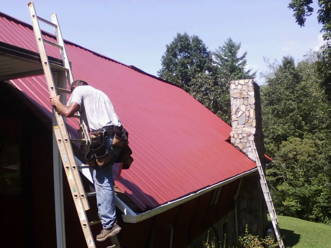 Roofer on a ladder beside a red metal roof and stone chimney — Soffit & Fascia Repair by Roofing Brothers in Line Lexington